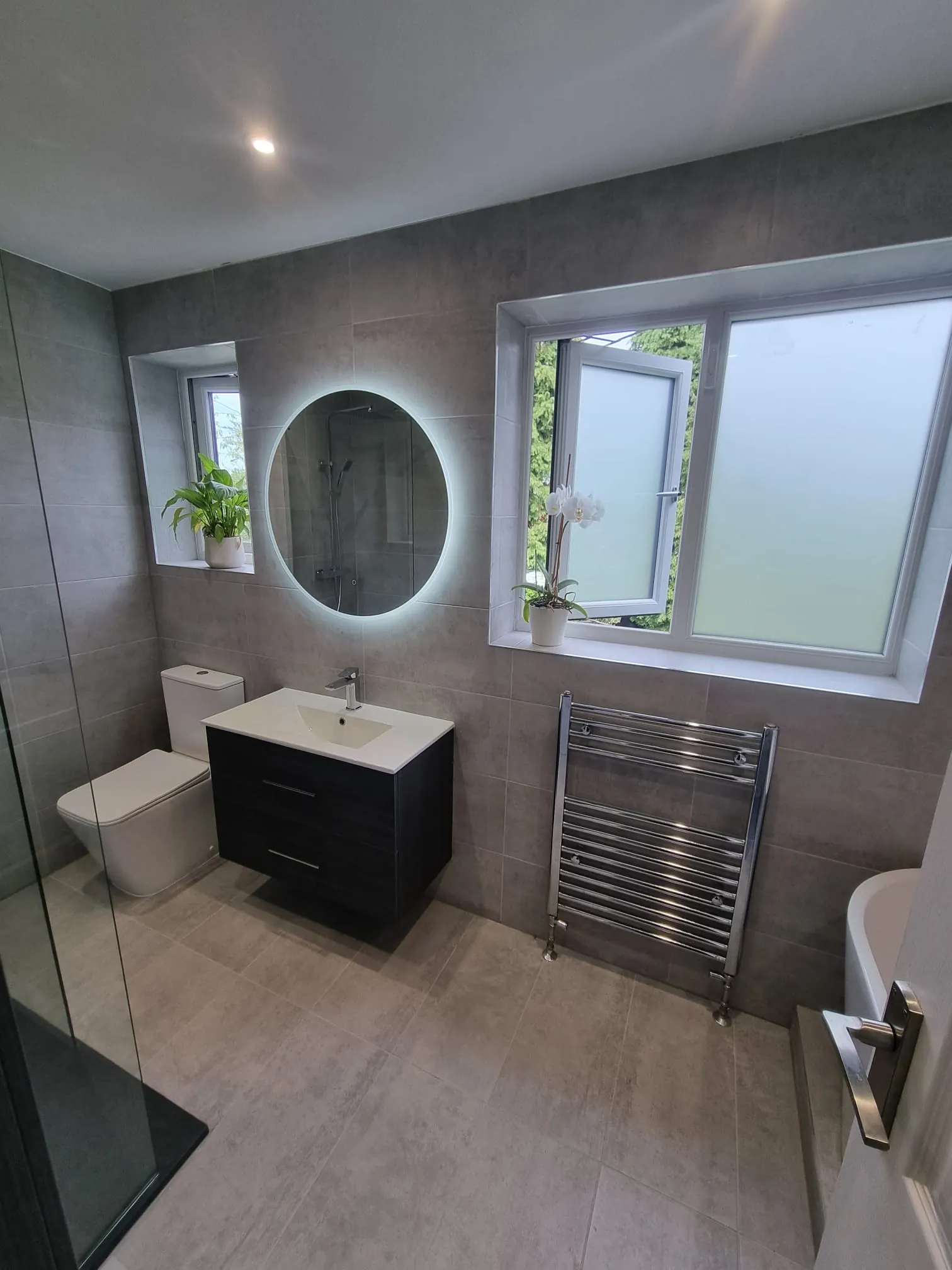 Grey tiled bathroom with a black vanity, round illuminated mirror and chrome towel rail