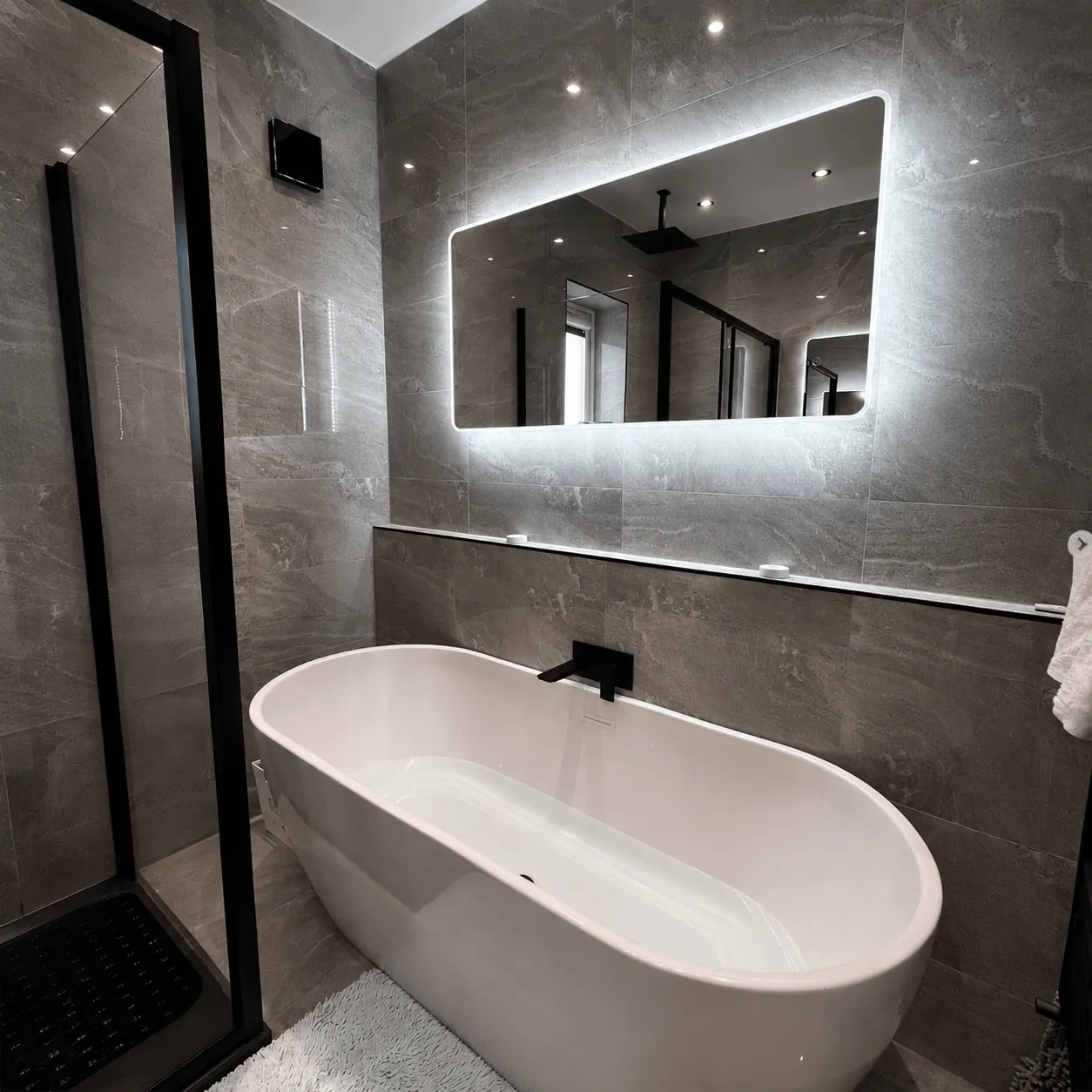 Freestanding bath beneath a wide illuminated mirror in a grey bathroom