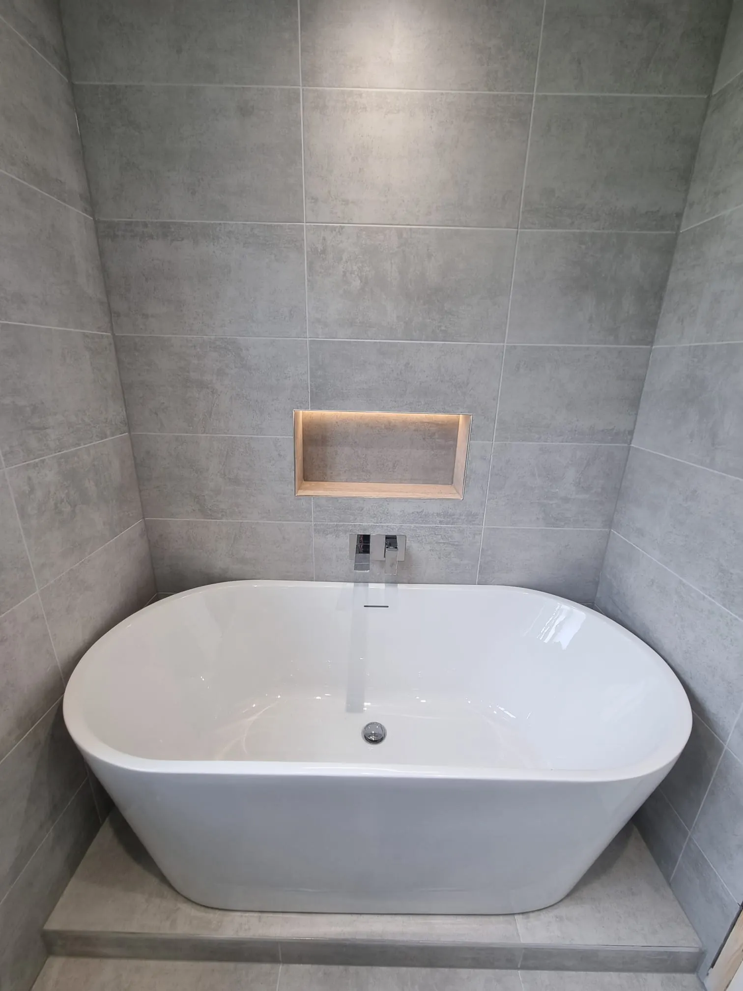 Freestanding oval bath beneath an illuminated recess in a grey tiled bathroom