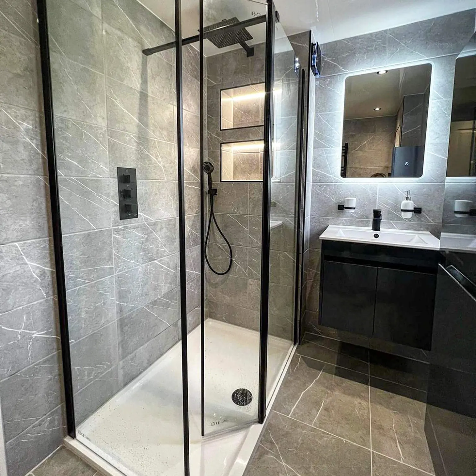 Grey tiled ensuite with a glass shower enclosure, black vanity and illuminated shelves